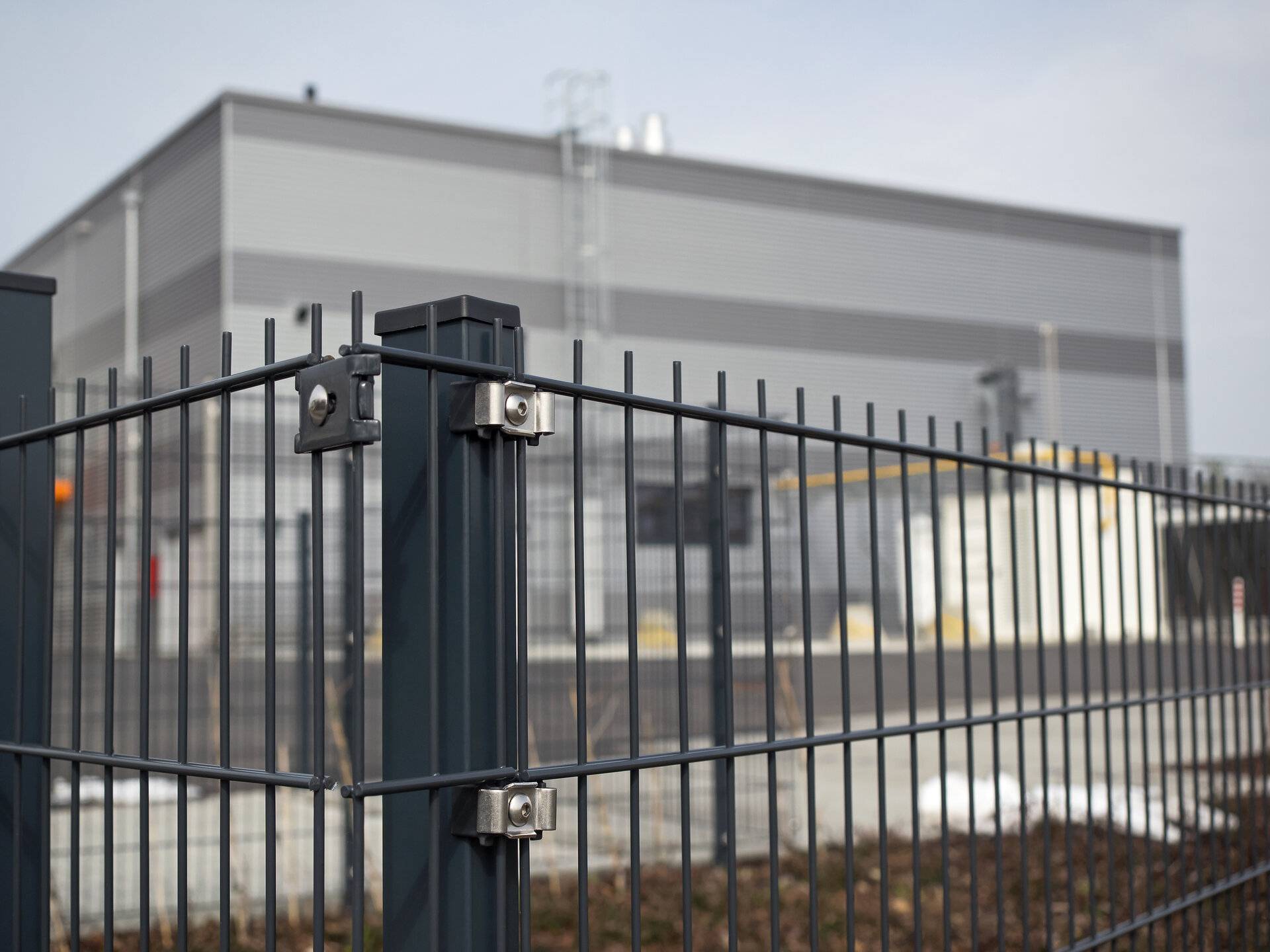 Blick auf einen Metall-Gitter-Zaun (Doppelstabmattenzaun), im Hintergrund hochmoderner Industrie- und Forschungsbetrieb (High-Tech-Unternehmen der Automobilzulieferindustrie), Fokus auf Stahlpfosten im Vordergrund.